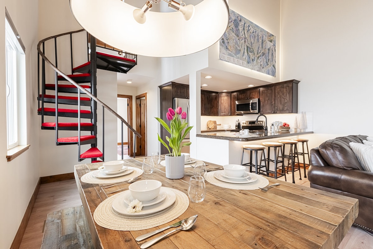 A dining area presents a wooden table set for six, featuring white dishware and a vase of tulips at its center. The open layout reveals a spiral staircase leading to the upper level, with modern kitchen amenities visible in the background.
