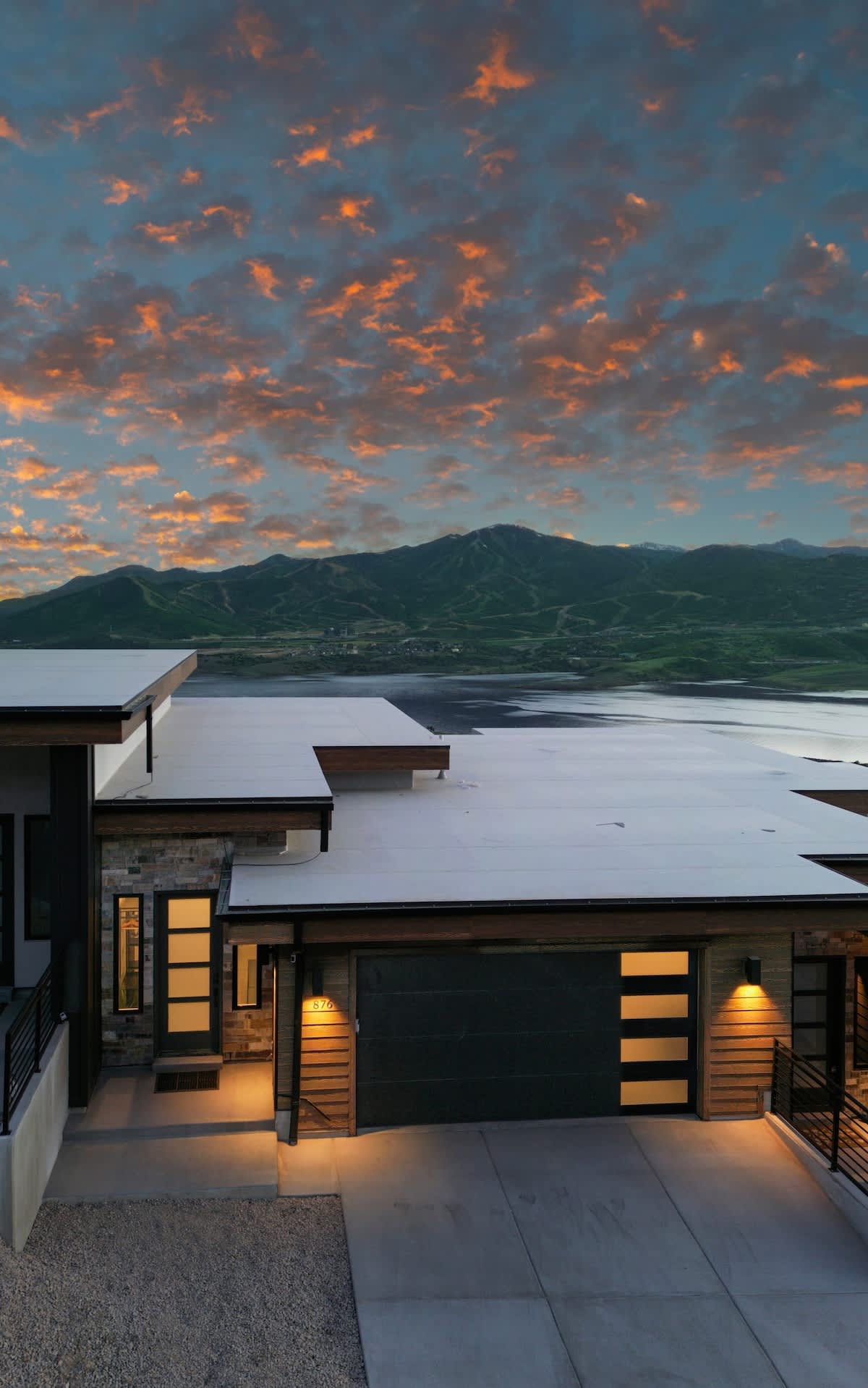 A modern home exterior is highlighted against a vibrant sunset sky, showcasing a blend of architectural lines and natural scenery. The garage door and entryway lights illuminate the pathway, while the expansive view of the mountains and reservoir stretches beyond.