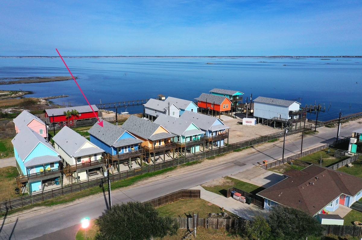 An aerial view captures a waterfront community featuring colorful homes along Laguna Madre Bay. The scene showcases a row of elevated structures with varied hues, surrounded by scenic water views and lush greenery, emphasizing the coastal setting.