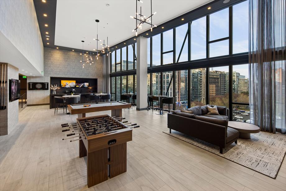 The open communal lounge showcases large windows that illuminate the space. Comfortable seating is arranged near a foosball table, and a kitchen area is visible in the background. Stylish lighting fixtures enhance the modern aesthetic, while views of the city skyline are presented through the glass walls.