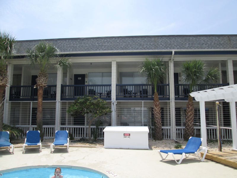 The image displays a two-story building featuring several balconies, each with black railing. Palm trees are visible on either side, adding to the tropical ambiance. A pool with blue lounge chairs is located in the foreground, suggesting a relaxed outdoor atmosphere.