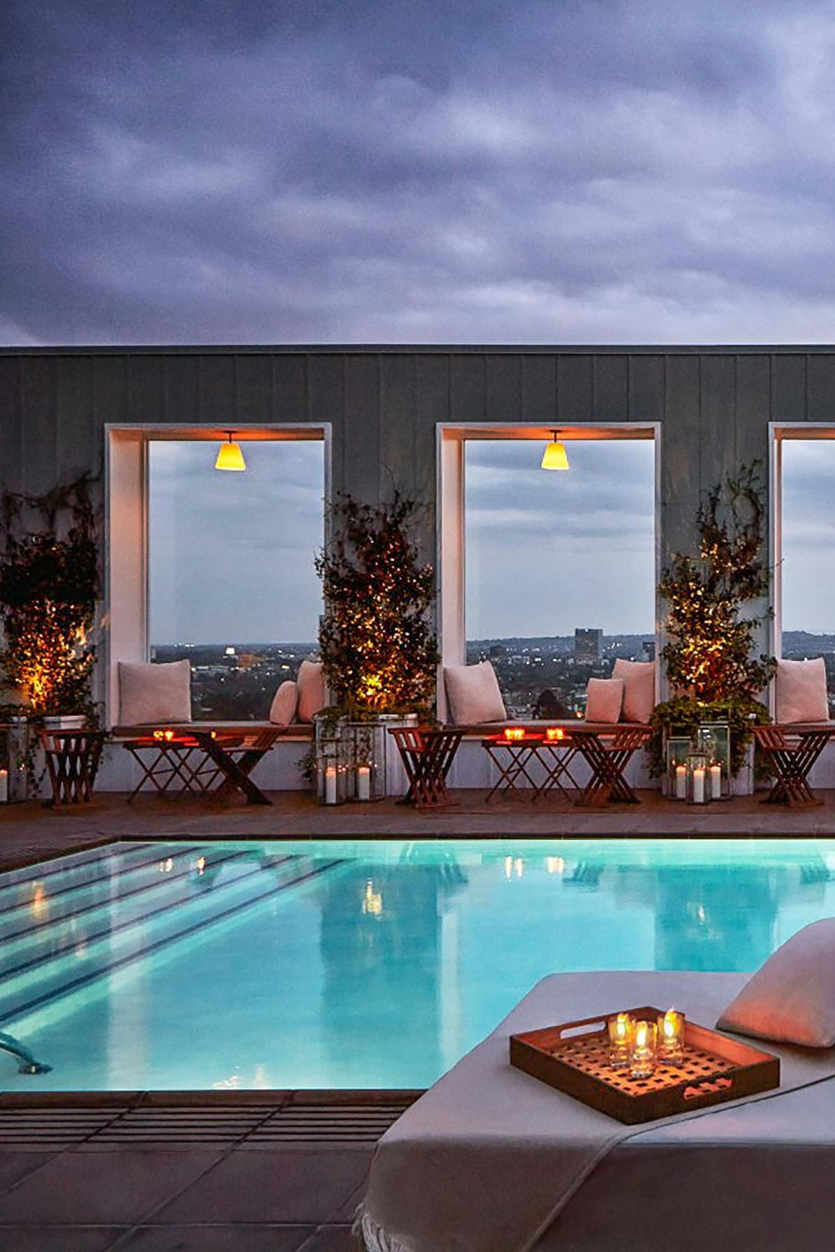 An elegant rooftop pool area is surrounded by floor-to-ceiling windows. Soft lighting and potted greenery create a serene ambiance. Lounge chairs are positioned beside the shimmering water, offering a tranquil space for relaxation. A tray with candles can be seen on the edge of a nearby chaise.