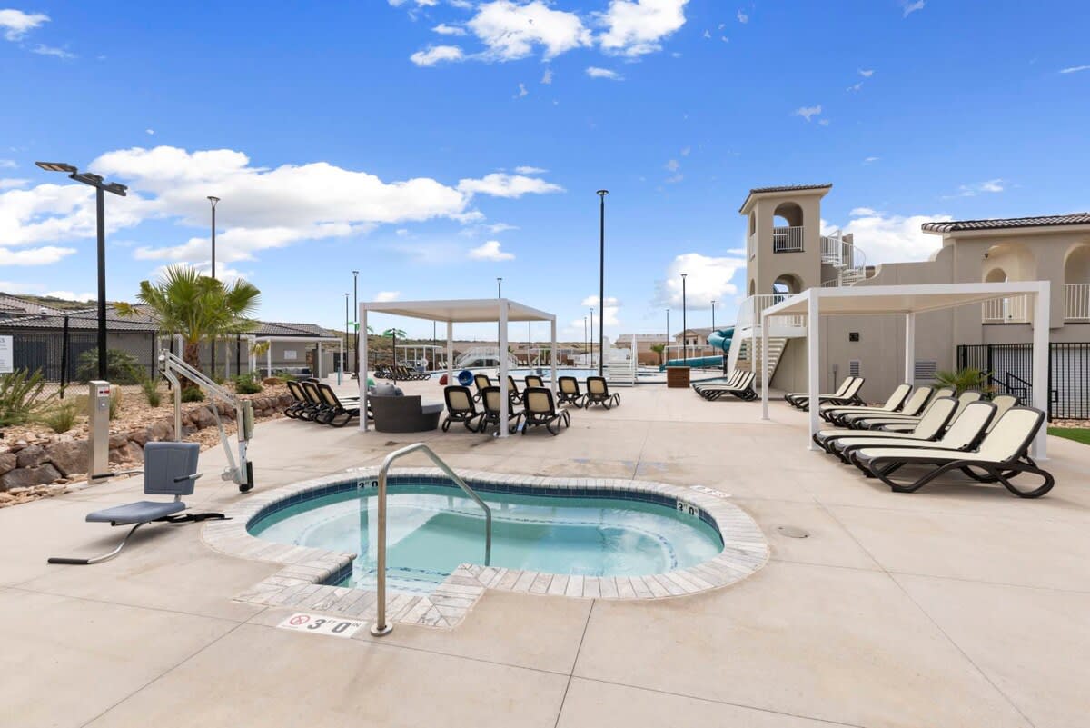 An outdoor swimming pool area features a hot tub surrounded by paved surfaces and lounge chairs. Shade structures are positioned throughout, offering a respite from the sun. Palm trees and lighting enhance the inviting atmosphere of this communal retreat.