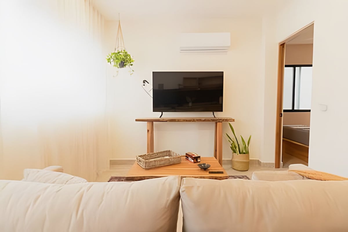 A comfortable living area is featured, showcasing a mounted television on a wooden stand, accompanied by a decorative plant. Natural light filters through sheer curtains, illuminating a cozy sofa and a coffee table with decorative items.