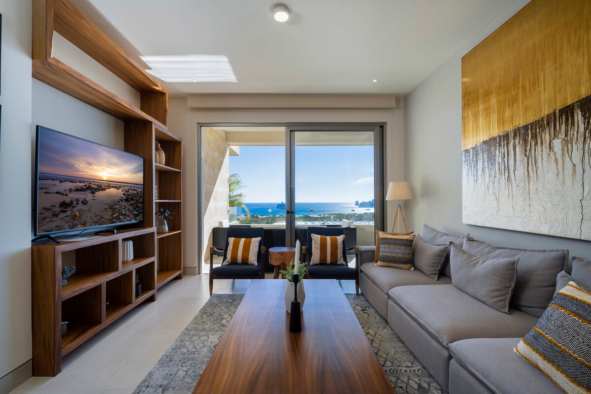 A modern living room is presented with a comfortable sofa and two armchairs, positioned around a wooden coffee table. A large television is mounted on the wall. Floor-to-ceiling sliding glass doors open to a private balcony with ocean views. Natural light fills the space.