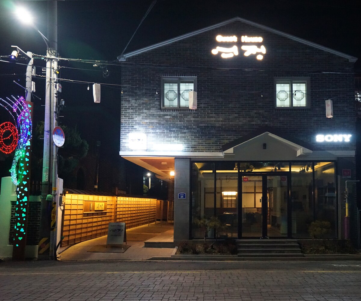 The exterior of the guesthouse is shown at night, featuring modern brick architecture. A well-lit entrance is visible, with welcoming signage. Decorative lights adorn the outdoor space, adding ambiance. The clean, clear path leads to the entrance, framed by greenery on either side.