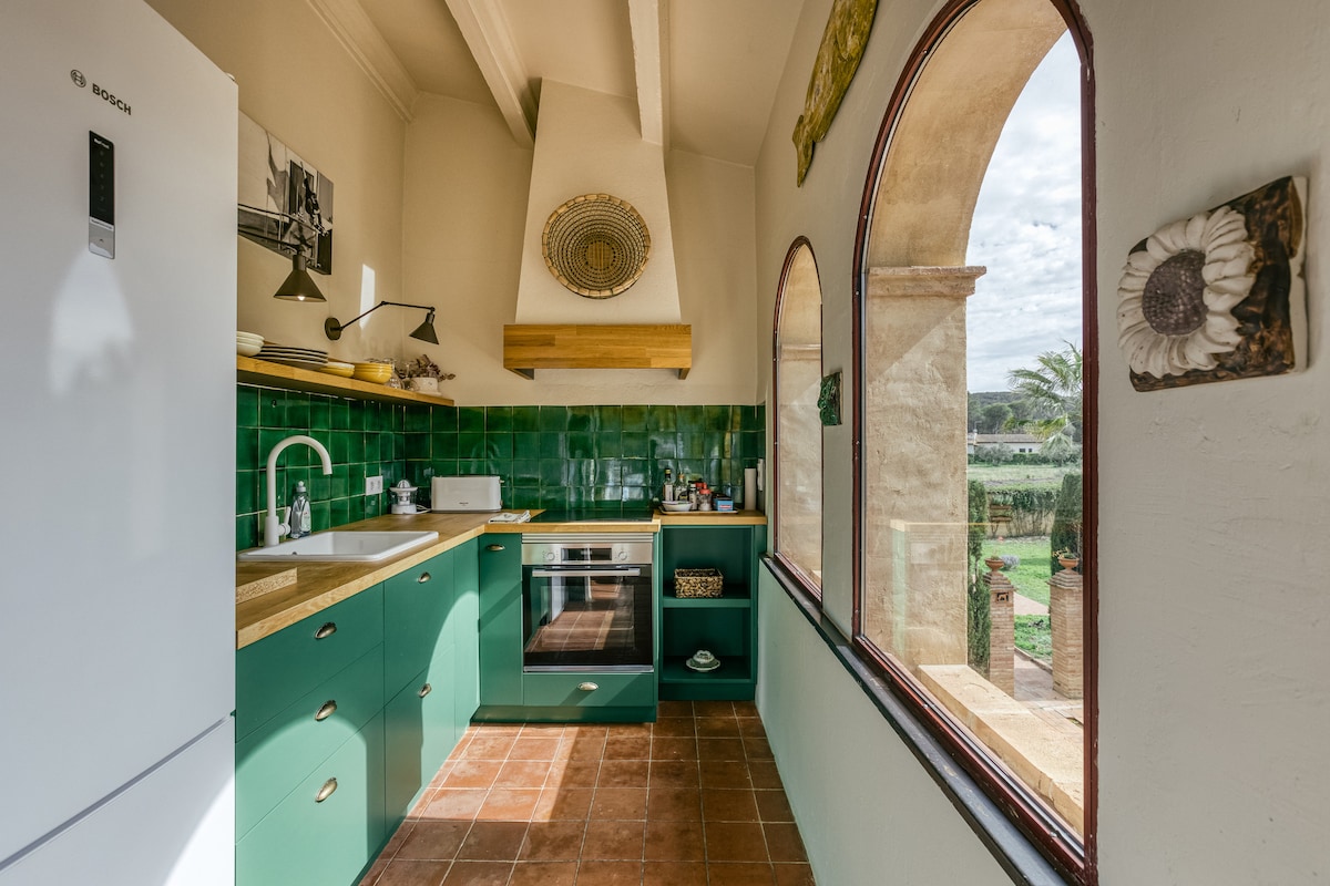 The kitchen features a modern design with teal cabinetry and white countertops. A large window arches over the sink, providing natural light and a view of the garden. Stainless steel appliances, including an oven, complete the functional space, enhancing the culinary experience.