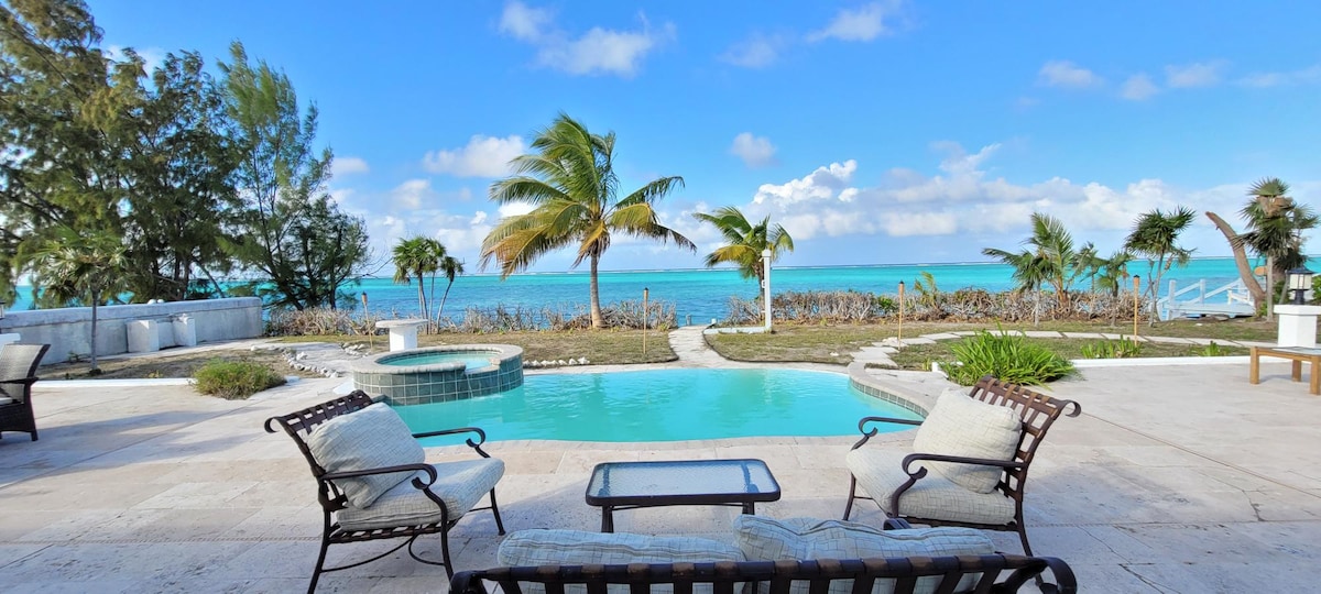 A landscaped outdoor space is highlighted by a crystal-clear pool, framed by lounge chairs and a small table. Lush palm trees sway gently in the background, providing tropical ambiance, while the serene turquoise water of the beach stretches beyond the property.