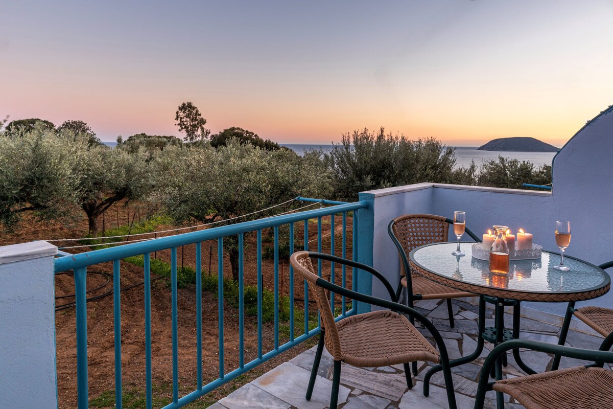 A serene outdoor balcony is depicted, featuring a round glass table set with glasses and flickering candles. Olive trees are visible in the background, while the horizon showcases a peaceful sunset over the sea.