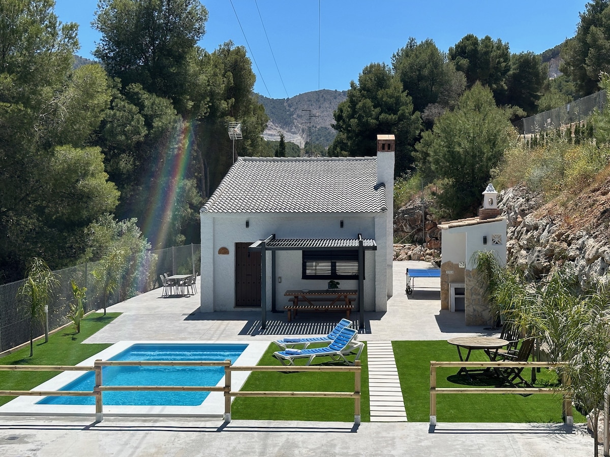A spacious outdoor area features a private pool surrounded by green grass. Lounge chairs are positioned near the pool, and a barbecue area is visible nearby. The house is framed by trees and mountains, providing a serene backdrop.