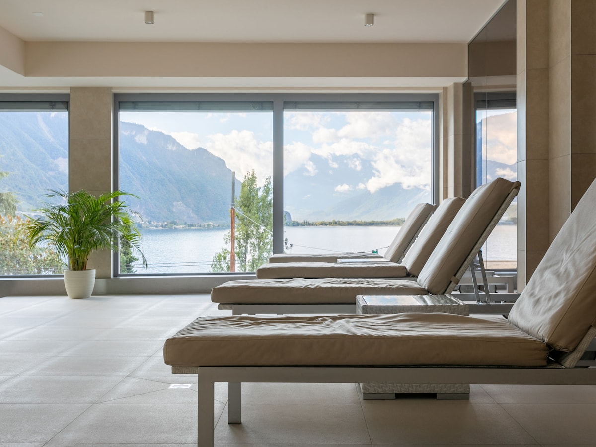 The indoor relaxation area features several sun loungers with soft beige cushions. Large floor-to-ceiling windows provide unobstructed views of the lake and surrounding mountains, allowing natural light to illuminate the space. A potted plant adds a touch of greenery to the serene environment.