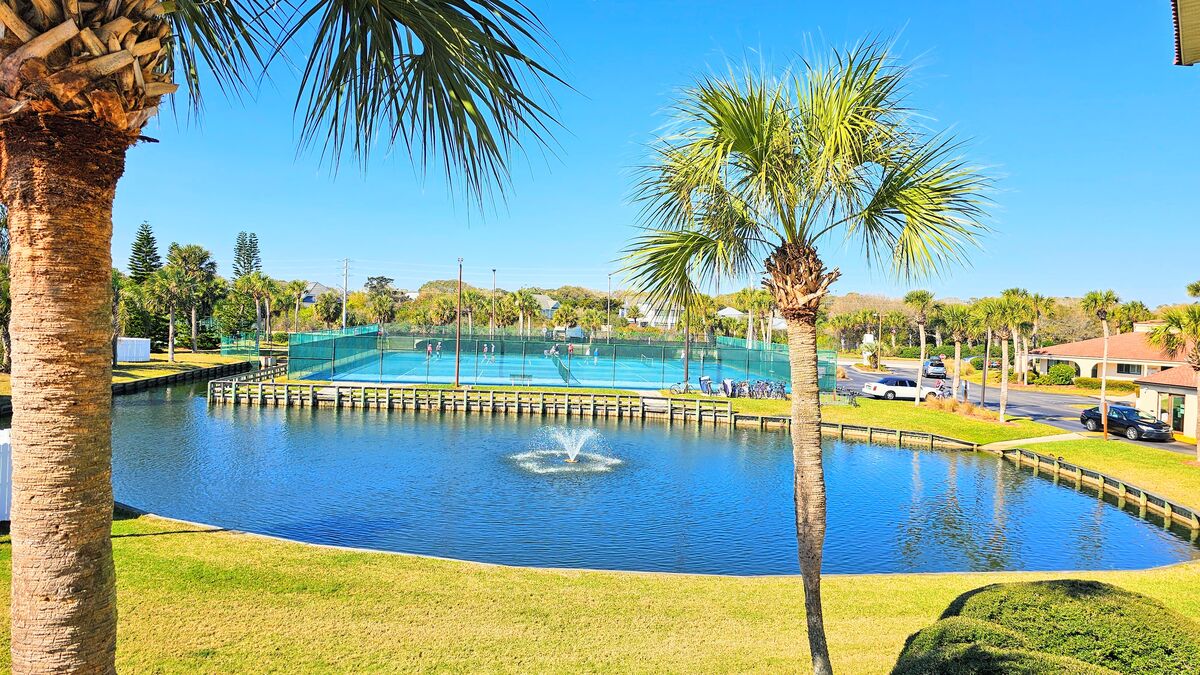 A serene view showcases a catch-and-release pond with a fountain, surrounded by well-manicured greenery. In the background, four tennis courts are visible, framed by palm trees and a clear blue sky, contributing to a tranquil outdoor environment.