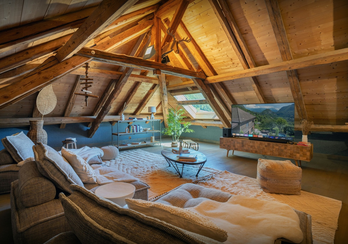 A spacious living area features sloped wooden ceilings and exposed beams. Comfortable sofas are arranged around a central coffee table, with a large television positioned against the wall. Natural light fills the room, highlighting the textured decor and soft area rug.