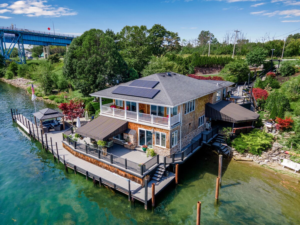 A premier waterfront boutique bed and breakfast is situated surrounded by lush greenery and water. The multi-level structure features an inviting deck, with outdoor seating areas and vibrant landscaping. Solar panels are visible on the roof, contributing to the eco-friendly design.