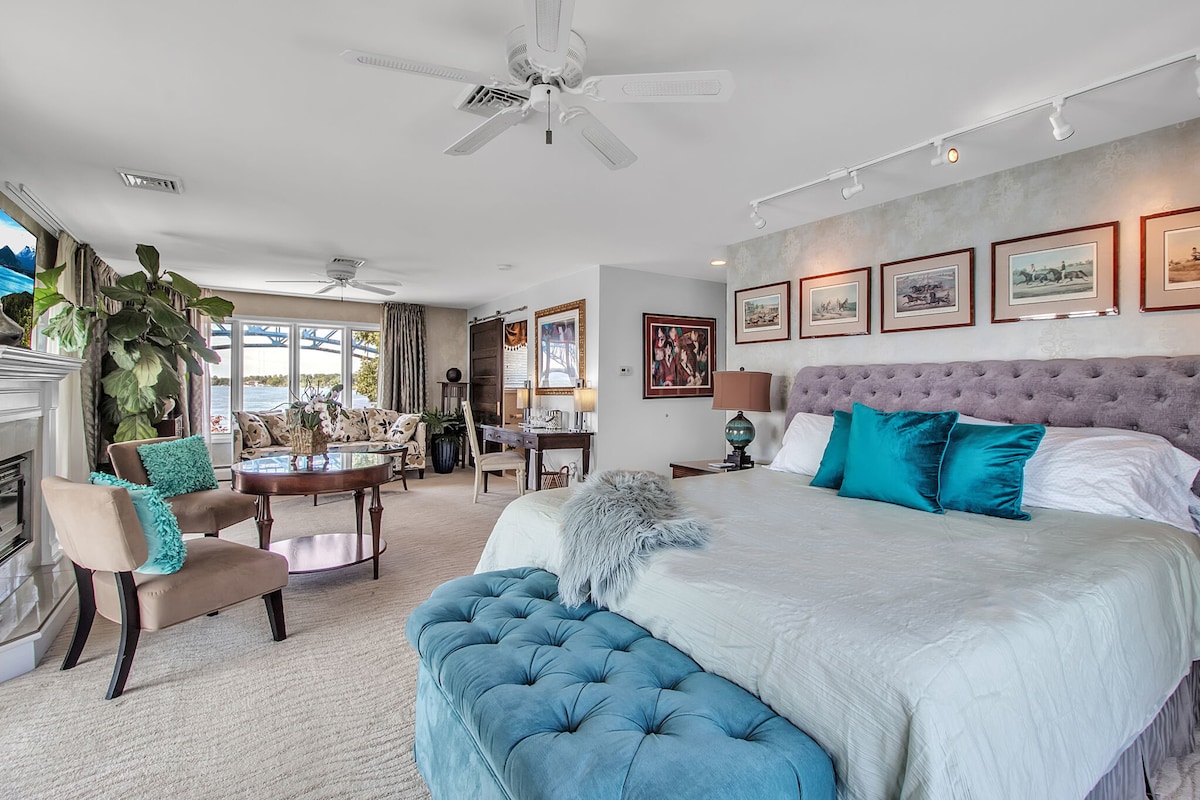 A spacious bedroom is furnished with a plush king bed adorned with decorative pillows, complemented by a stylish tufted bench at the foot. Natural light pours in through large windows, illuminating a cozy seating area with floral-patterned chairs and a dining table. A soft, neutral palette enhances the relaxing ambiance.