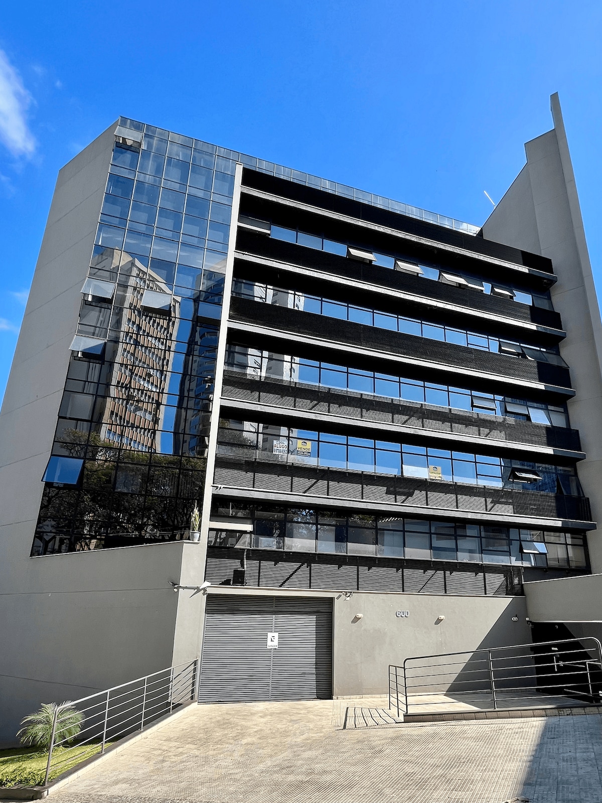 A modern multi-story building is depicted, featuring a sleek design with extensive glass windows reflecting the surrounding cityscape. The entrance is accessible via a ramp, and the structure is framed by a clear blue sky.