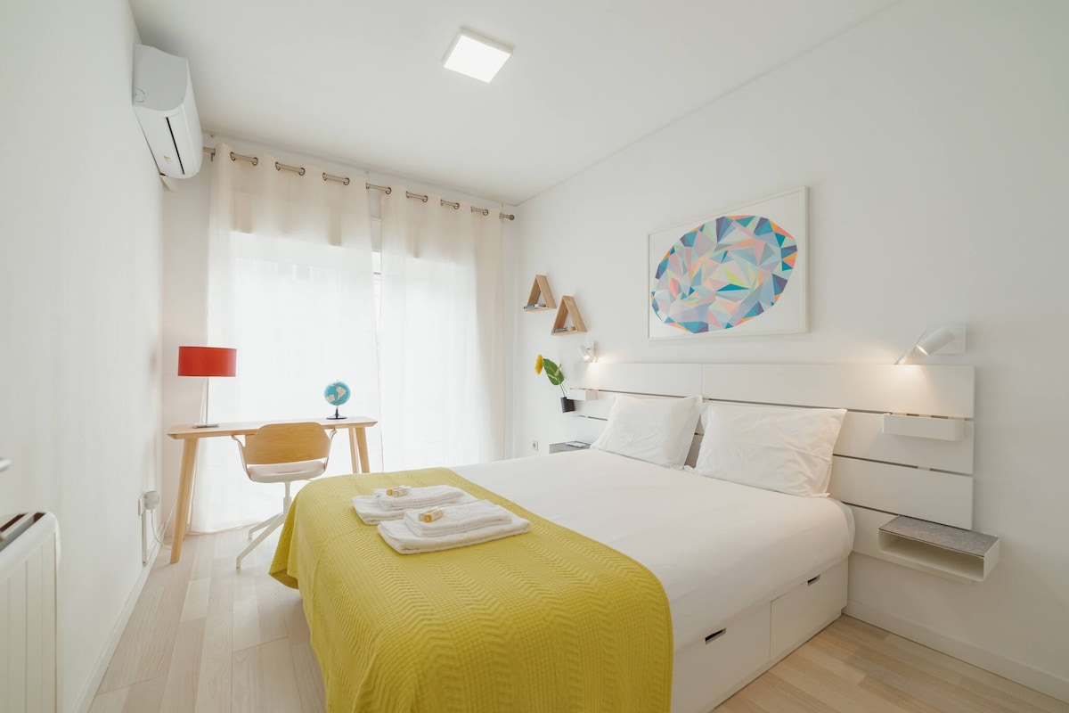A bright bedroom features a spacious double bed with fresh linens and a yellow blanket. A desk with a lamp and decorative elements enhances the ambiance. Natural light filters through sheer curtains, creating a warm atmosphere, while modern wall art adds a touch of color.