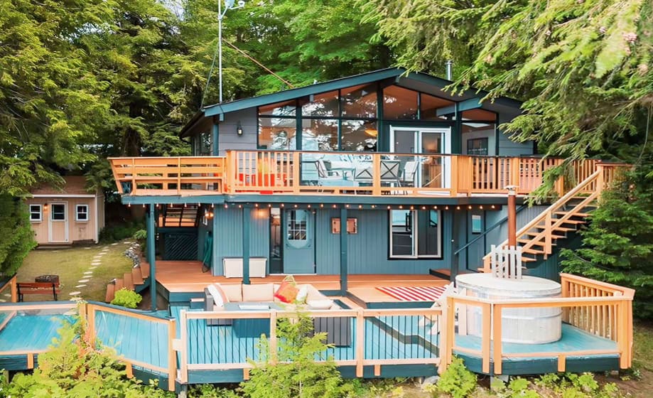 The exterior of the cottage showcases a modern design surrounded by lush greenery. A wraparound deck features seating areas and a hot tub. Large windows bring in natural light, and a staircase leads to an upper deck, offering elevated views of the surrounding landscape.
