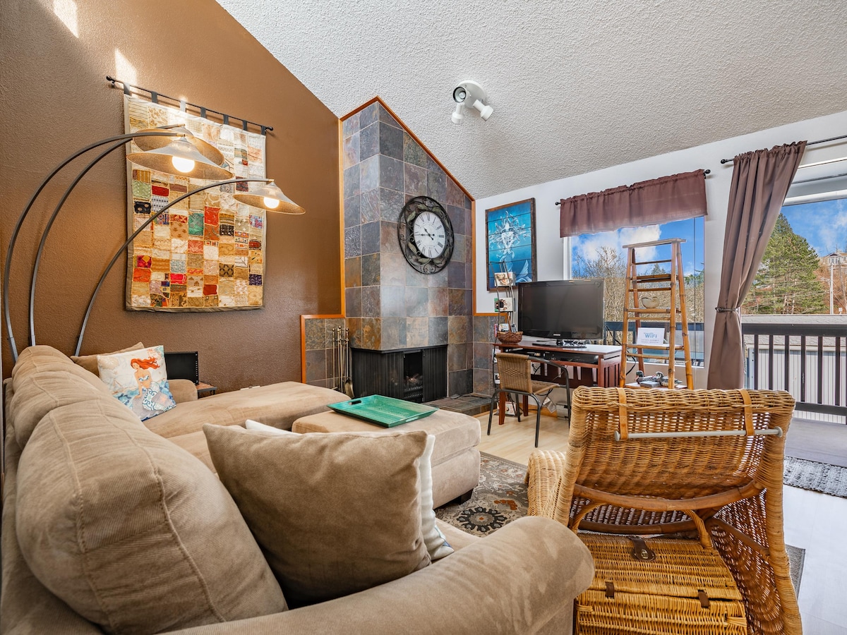 A cozy living area features a sectional sofa and a wicker chair, with a large clock mounted on the wall. A stone fireplace adds texture, and a TV is positioned nearby. Natural light flows in from the window, framed by dark curtains, enhancing the relaxed atmosphere.