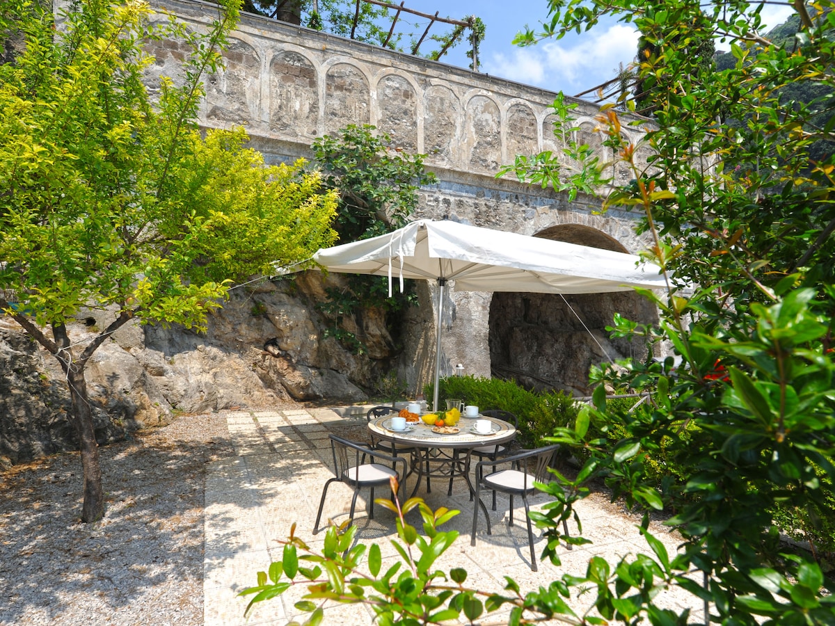 An outdoor dining area is surrounded by lush greenery and stone walls. A large table is set with refreshments and shaded by a canopy. The ground is paved with light stones, creating a tranquil atmosphere amidst the natural landscape.