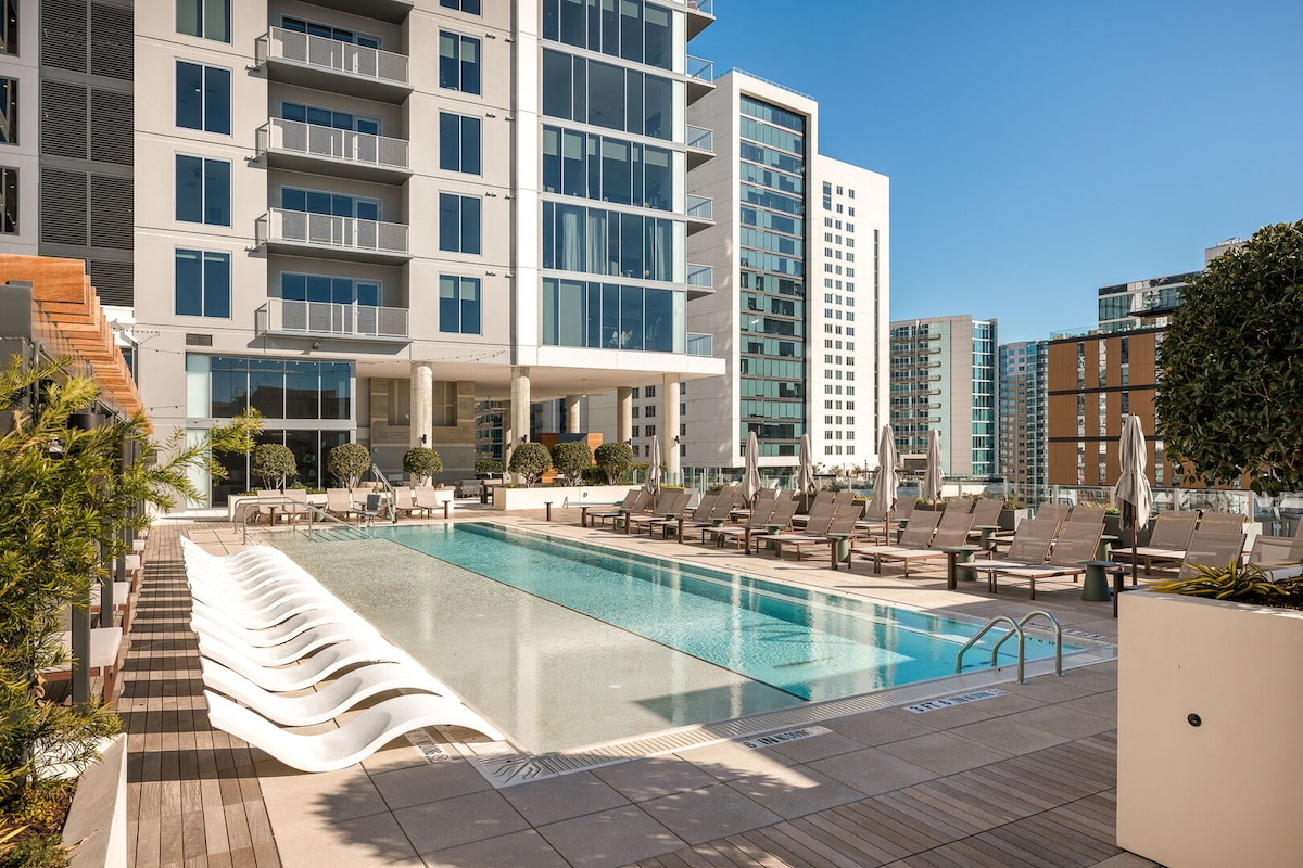 An inviting pool area is framed by modern architecture, featuring a sleek pool accompanied by lounge chairs arranged neatly along the deck. Lush greenery enhances the outdoor space, complemented by sunny skies and city buildings in the background.