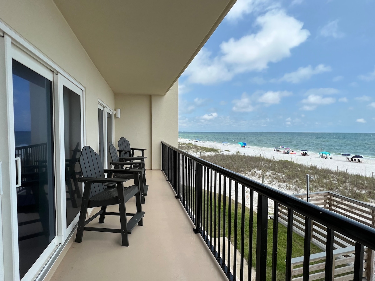 A spacious balcony is featured, furnished with two black wooden chairs facing the ocean. The turquoise waters and white sandy beach can be seen in the distance, with colorful umbrellas dotting the shoreline. Soft clouds drift across a bright blue sky.