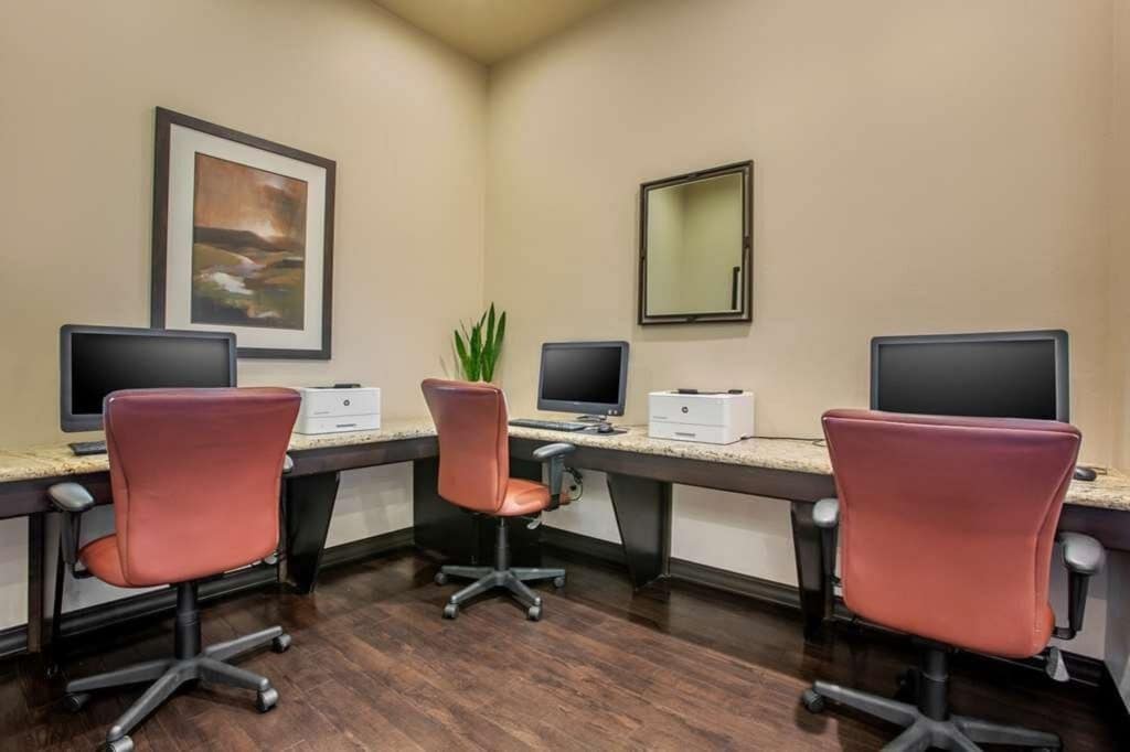 A workspace area is equipped with four computer stations, each paired with a rolling chair in a maroon hue. The dark wood floor contrasts with the light walls, and a mirror is mounted above the workspace. A plant adds a touch of greenery to the environment.