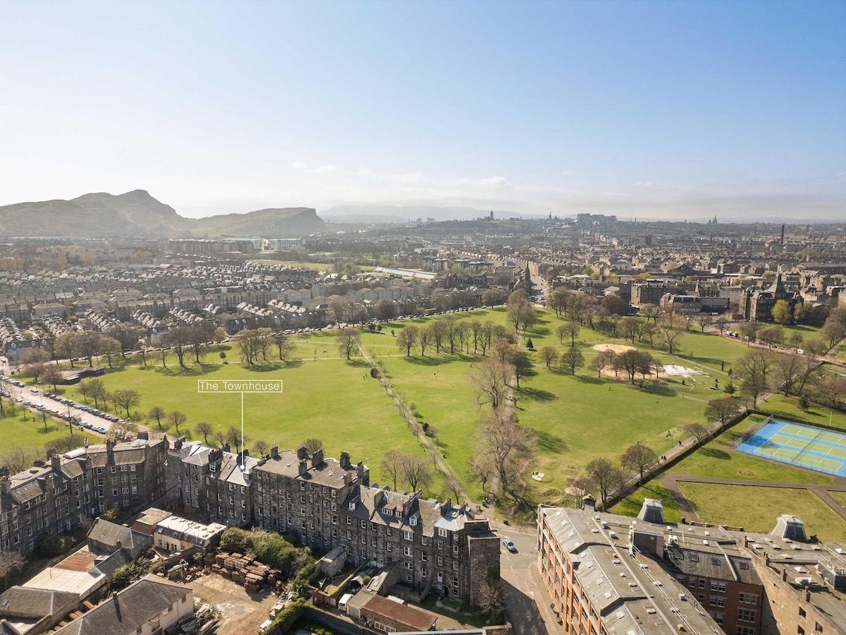 An aerial view showcases a long, historic Georgian townhouse framed by a public park. Lush greenery, pathways, and open spaces can be seen in the park area, while a scenic landscape of hills and city buildings creates a picturesque backdrop.