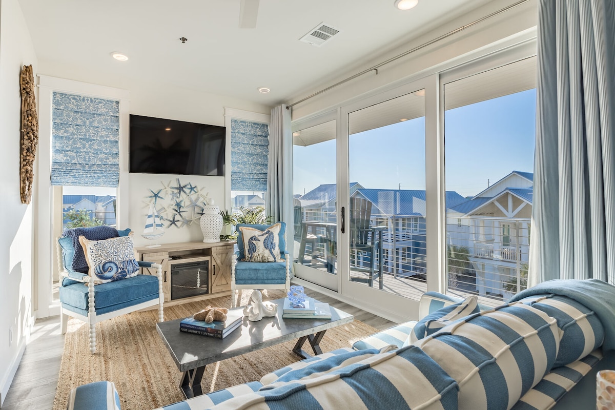 A bright living area features large glass doors that open to a scenic view. Comfortable seating is arranged around a coffee table, with coastal decor enhancing the space. Soft blue and white patterns on cushions complement the natural light streaming in.