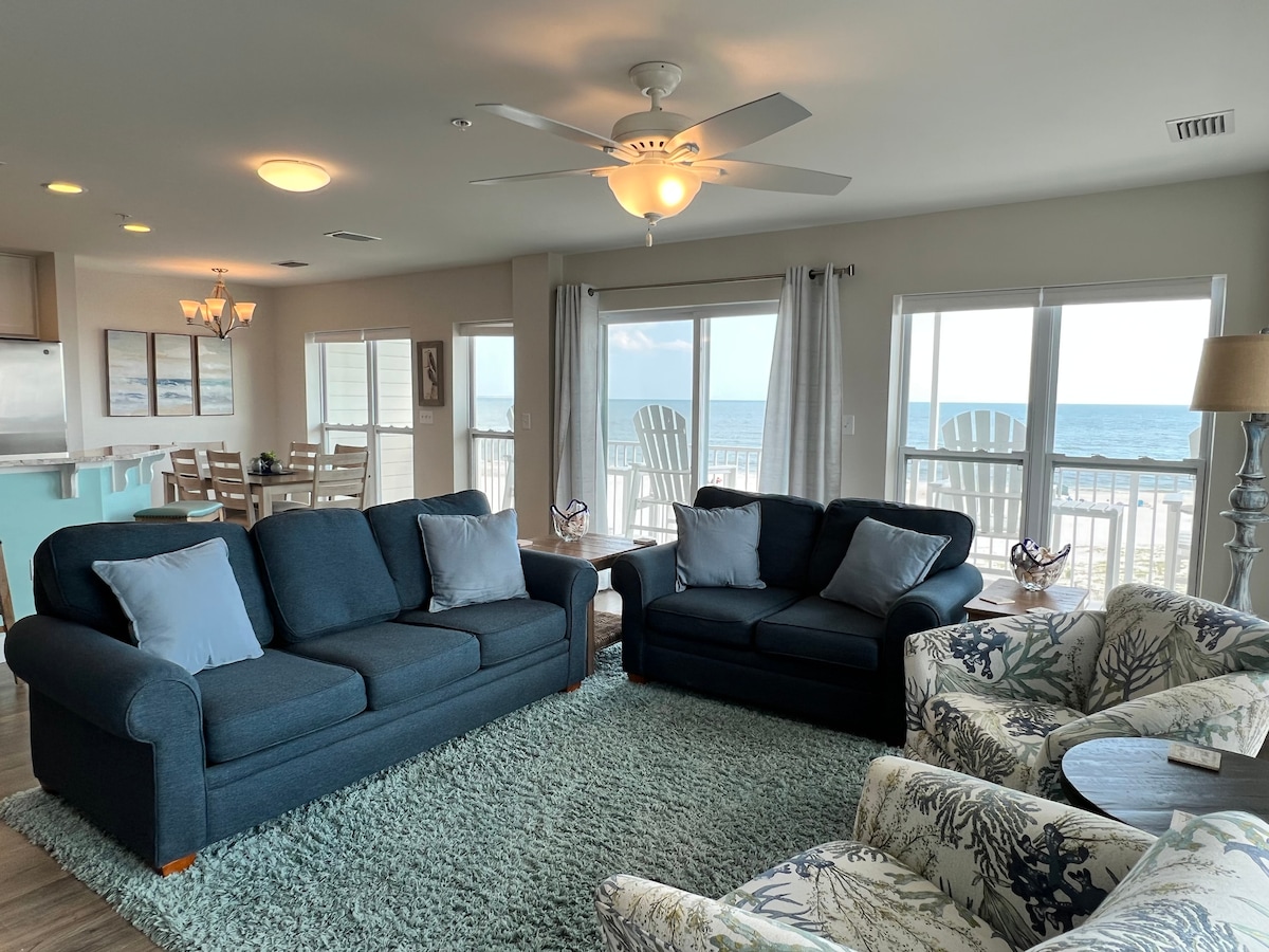 A comfortable living area features a blue sofa and armchairs, with decorative pillows enhancing the seating. Large windows offer scenic views of the gulf, while a rug adds texture to the clean floors. A dining table and kitchen are visible in the background.