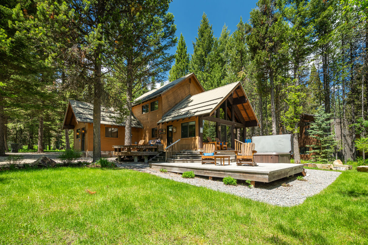 The exterior of Chandler's Lodge is set amidst a lush green landscape, featuring a spacious wooden deck and inviting outdoor seating. Surrounded by tall trees, the home showcases a combination of charming architectural elements and natural surroundings, creating a serene retreat atmosphere.