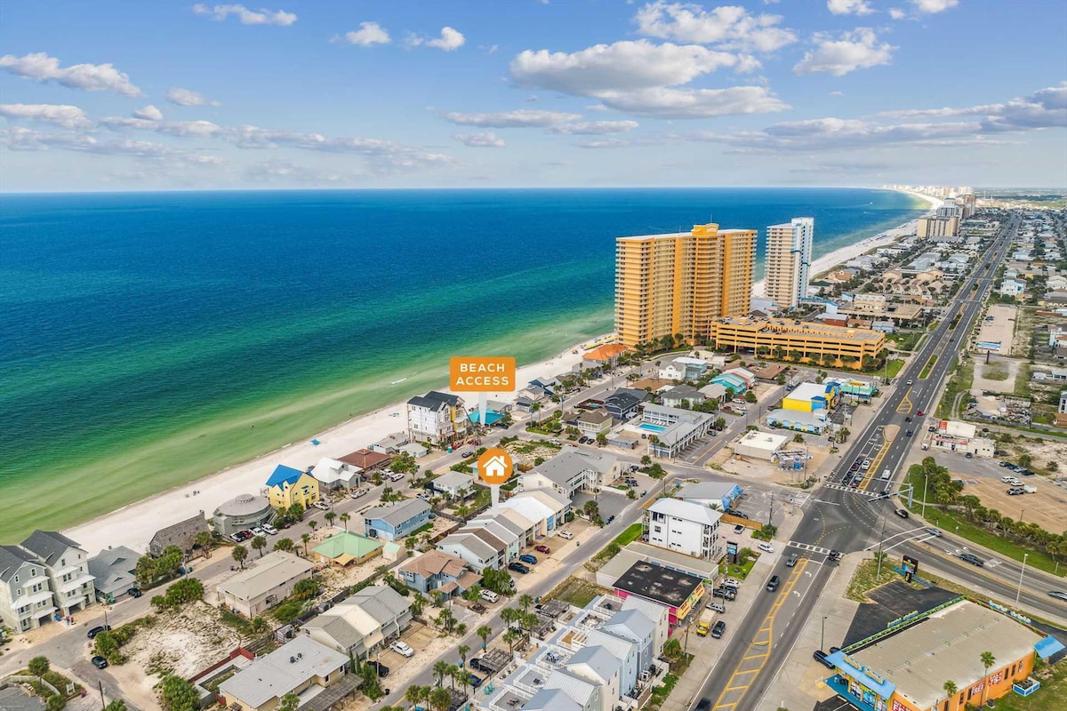An aerial view captures the turquoise waters and sandy beach, revealing the proximity to beach access. Coastal structures line the shore, with several buildings visible in the distance. The vibrant blue sky is scattered with fluffy clouds, highlighting the inviting landscape of the area.