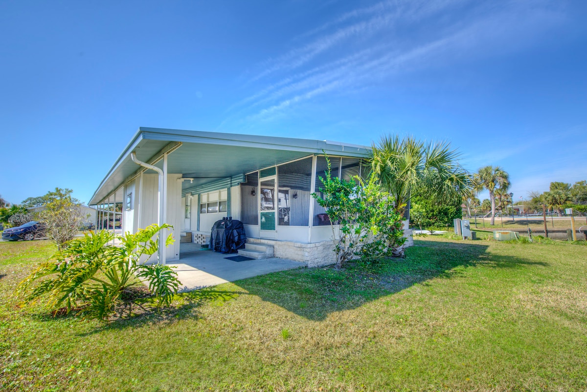 The exterior of a one-story residential home is shown, featuring a covered porch and a manicured lawn. Lush greenery and palm trees frame the property, while a clear blue sky is visible above, suggesting a bright and sunny day.