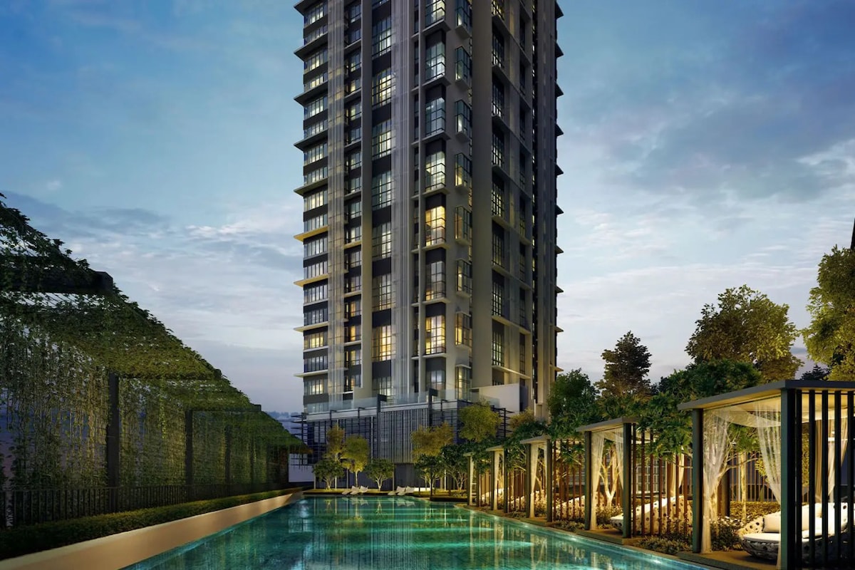 A modern high-rise building is captured at dusk, featuring sleek architectural lines and multiple levels of windows. The foreground includes a landscaped pool area with lounge cabanas and greenery, reflecting the building's illuminated exterior in the tranquil water.