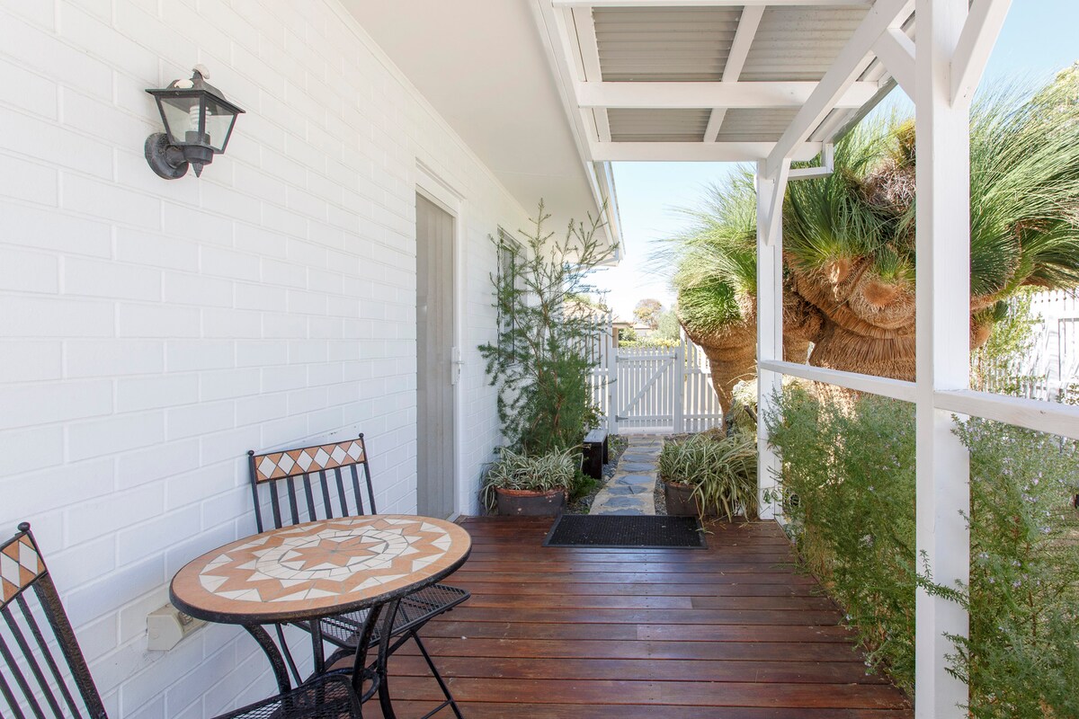 A small outdoor patio area features a round table with a decorative mosaic top and two black metal chairs. Lush greenery surrounds the space, enhancing the inviting atmosphere. A light fixture is positioned on the white wall near the entrance, providing subtle illumination.