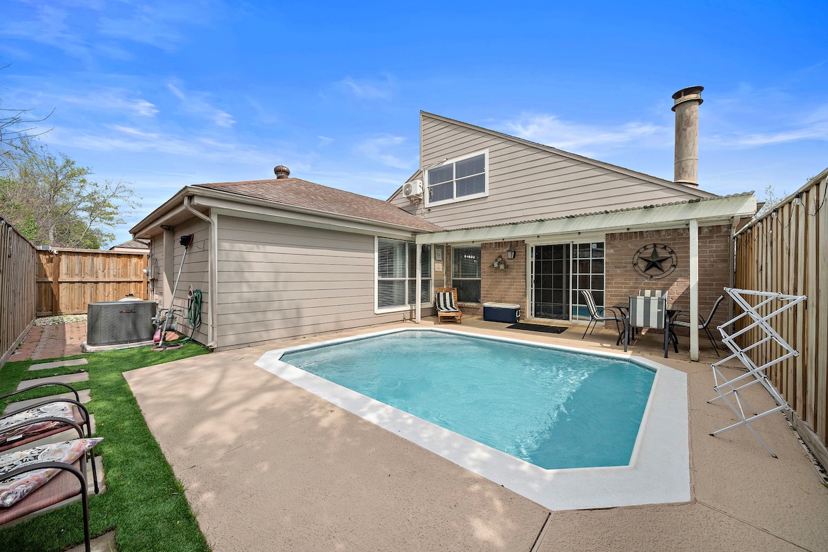 An inviting outdoor space features a private pool bordered by a light-colored patio. Comfortable seating options are arranged nearby, while a lush grassy area adds greenery. The exterior of the home is modern with large windows, allowing for natural light and views of the pool.