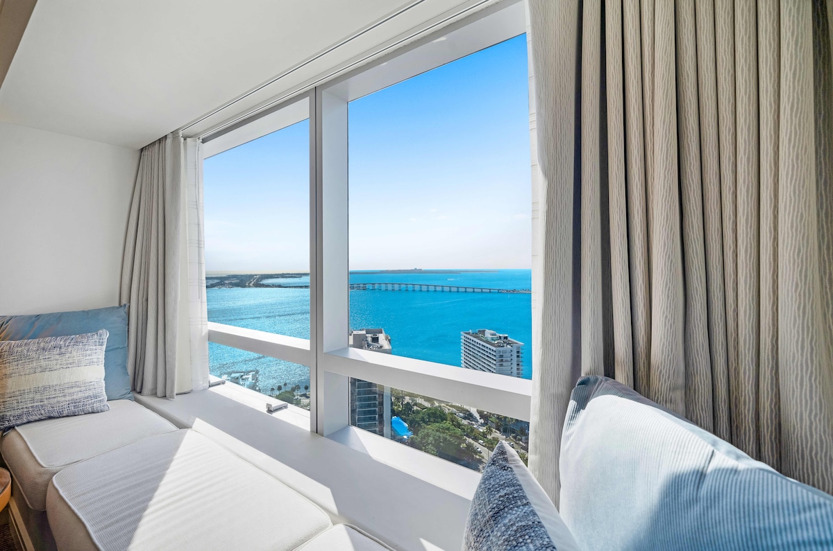 A comfortable seating area features a cozy nook with large floor-to-ceiling windows, offering stunning views of the sparkling blue ocean and distant horizon. Light-filtering curtains add softness while framing the picturesque scenery.