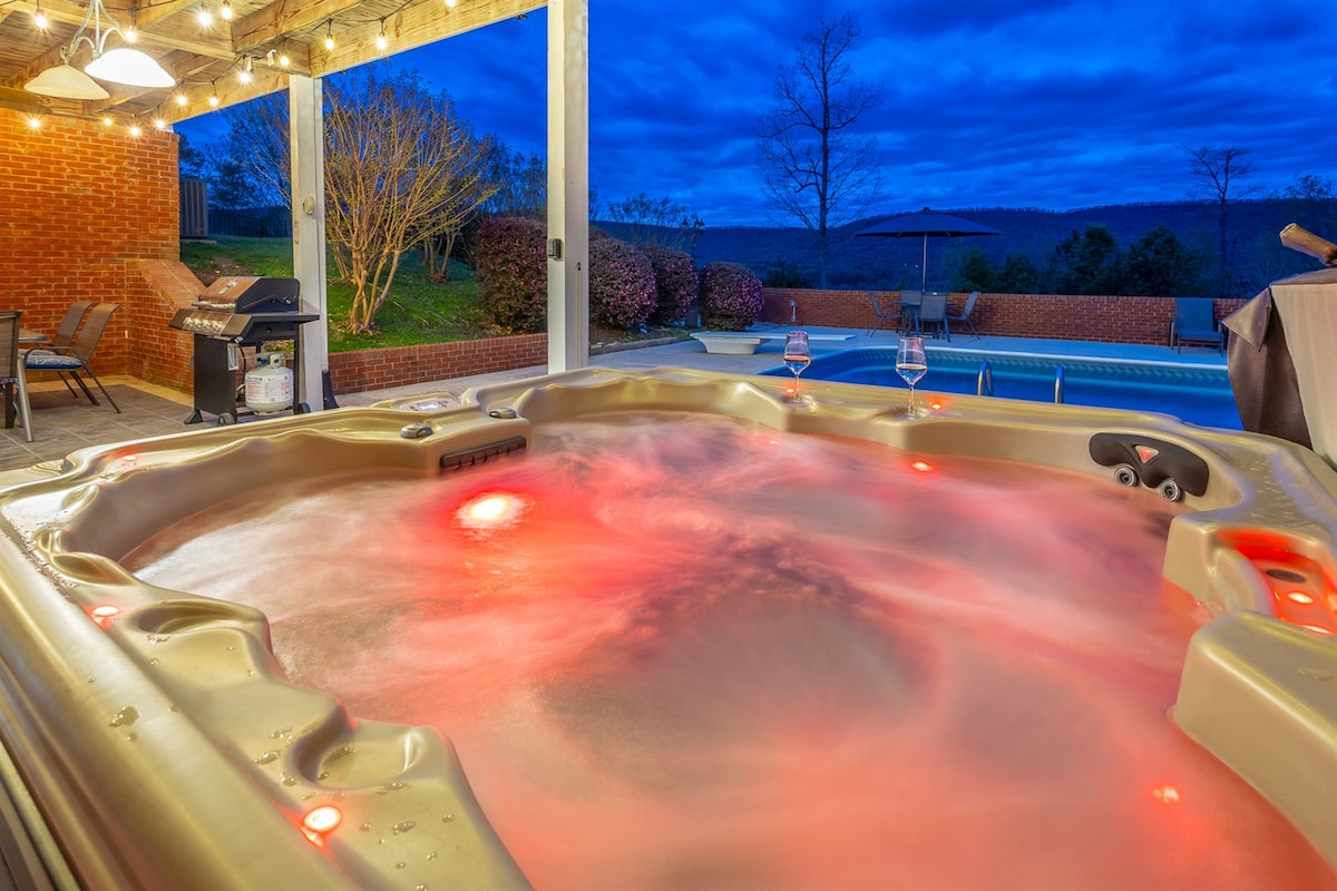 A hot tub is located on a covered patio, illuminated by soft lighting. The water appears inviting with a gentle swirl, and red lights add a calming glow. In the background, a swimming pool is visible, surrounded by greenery under a twilight sky.