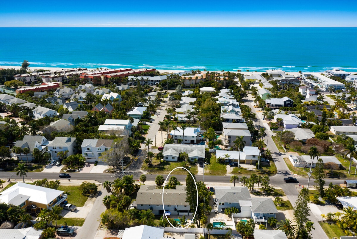 An aerial view showcases the elegant neighborhood with well-spaced homes surrounded by lush greenery. The shimmering Gulf waters are visible in the distance, indicating proximity to the beach, just a short walk away.