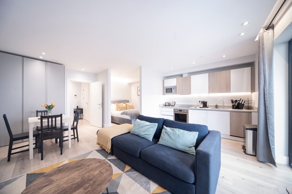 An open-plan living area is highlighted, featuring a comfortable sofa with accent cushions and a wooden coffee table. A dining table with four chairs is positioned nearby. The kitchen is visible with modern cabinetry and appliances, while a spacious double bed is situated in the background.