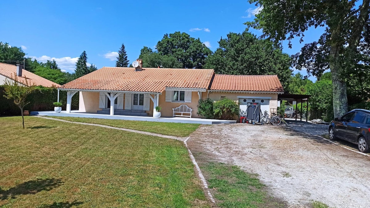 The villa features a single-story exterior with a tiled roof and covered porch areas. A large lawn area is visible in front, bordered by trees. Parking spaces accommodate vehicles, and bicycles can be seen near the entrance, providing easy access to outdoor activities.