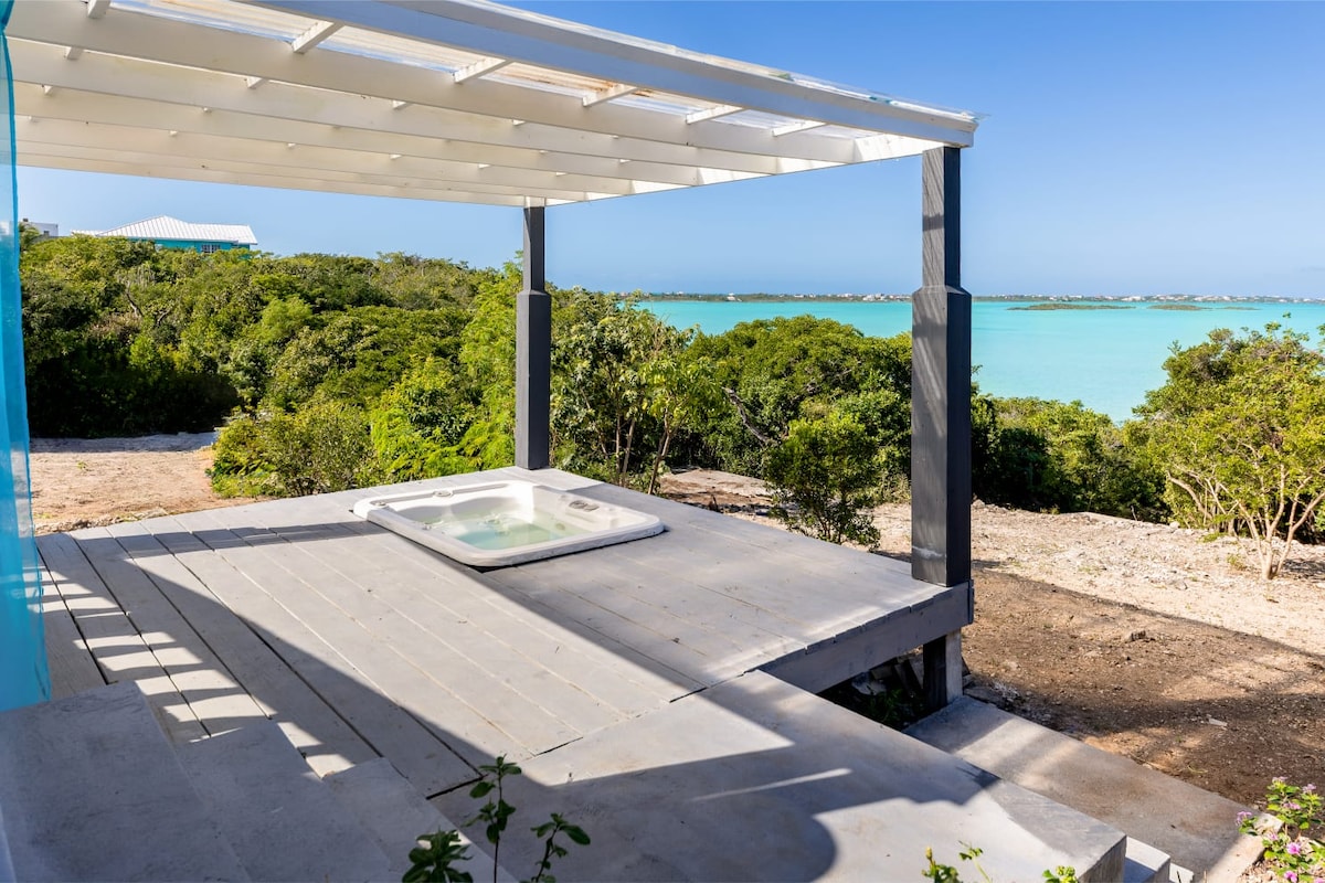 A shaded outdoor area features a whirlpool spa set on a wooden deck, with panoramic views of turquoise waters and lush greenery. The serene landscape offers a tranquil setting for relaxation, highlighted by the natural surroundings that enhance the peaceful ambiance.