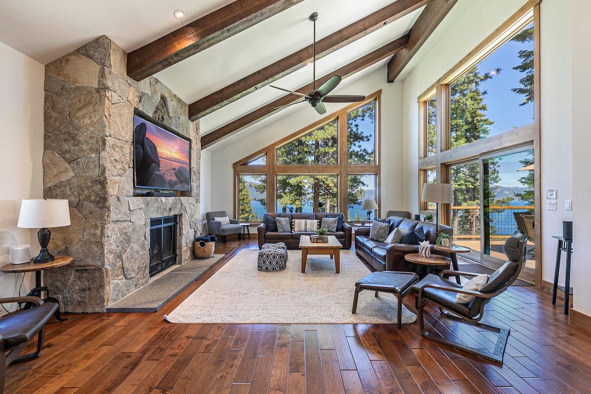 A bright living room features cathedral ceilings supported by wooden beams and a large stone fireplace. A wide screen TV is mounted on the wall, while ample seating is arranged on a plush area rug. Expansive windows provide views of the lake and surrounding trees.
