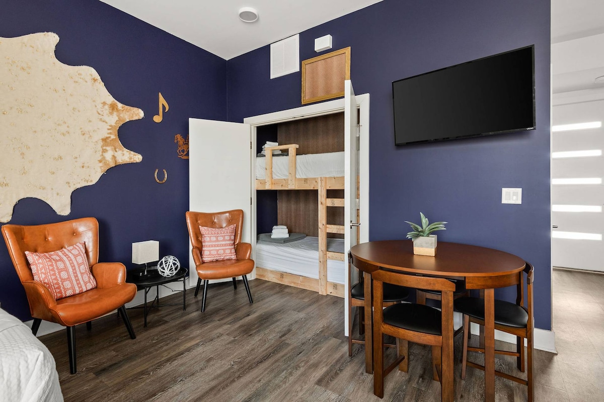 A compact studio space features a built-in bunk bed, a round table with four wooden chairs, and two orange armchairs with decorative cushions. A flat-screen TV is mounted on the wall, and a modern light fixture is visible near the entrance.