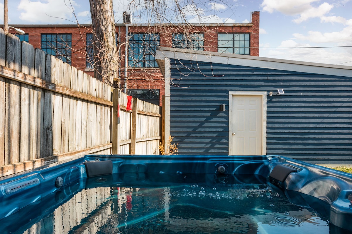 A private hot tub is set within a fenced backyard, offering a relaxing environment. The exterior of a blue structure is visible in the background, along with a wooden fence and trees, contributing to a tranquil outdoor setting.