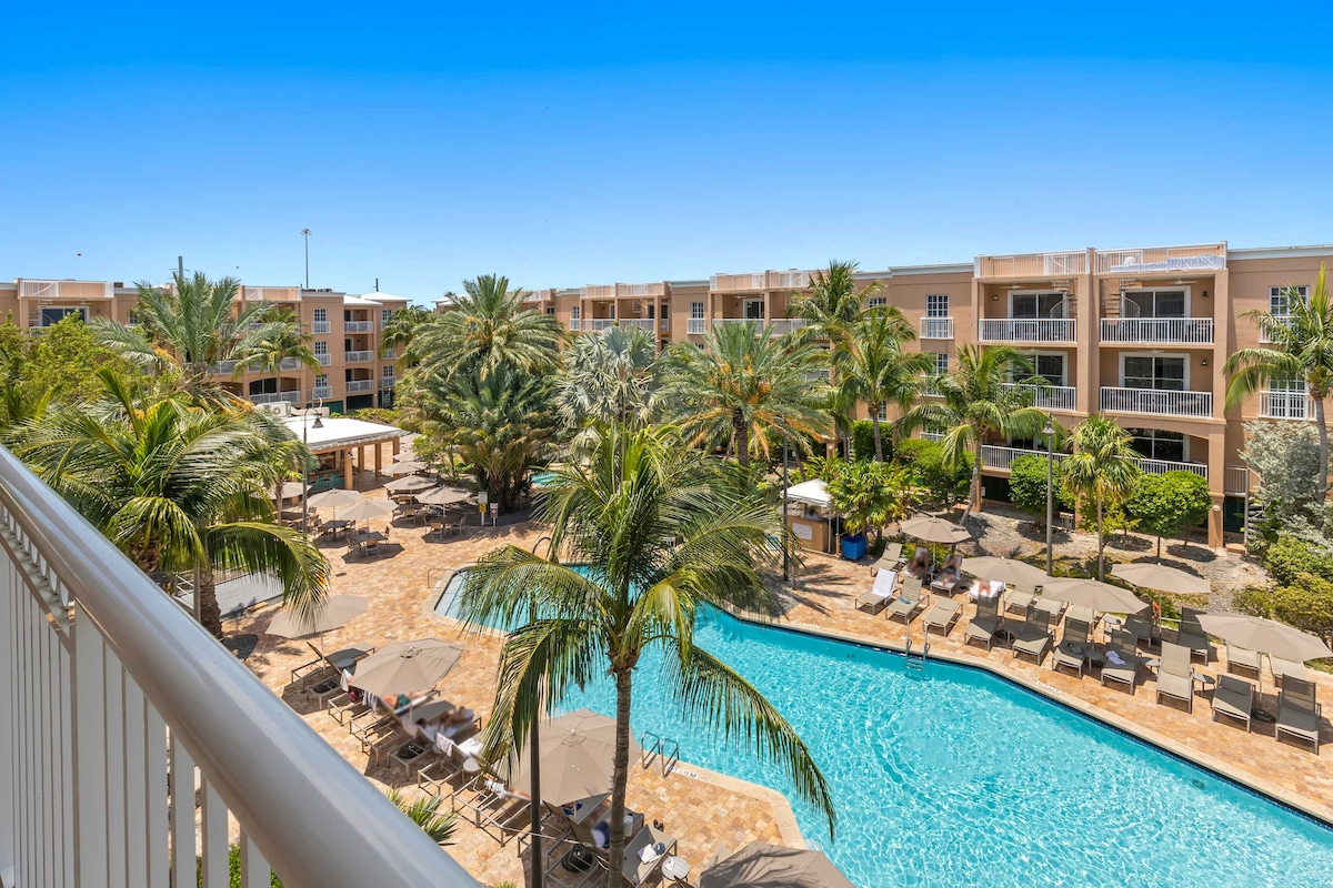 A view from a private balcony showcases the resort pool, surrounded by palm trees and a sun terrace. Lounge chairs are arranged neatly around the pool, while shaded seating areas provide options for relaxation. The bright blue sky enhances the atmosphere of leisure.