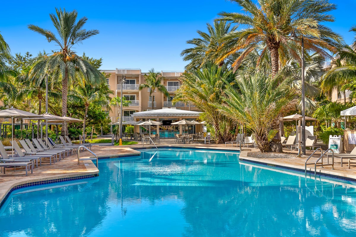 The beautiful resort pool is surrounded by tropical palm trees, creating a serene atmosphere. Loungers are positioned along the poolside, providing ample space for relaxation. The clear blue water reflects the sunny skies, inviting guests to unwind in this sunny oasis.