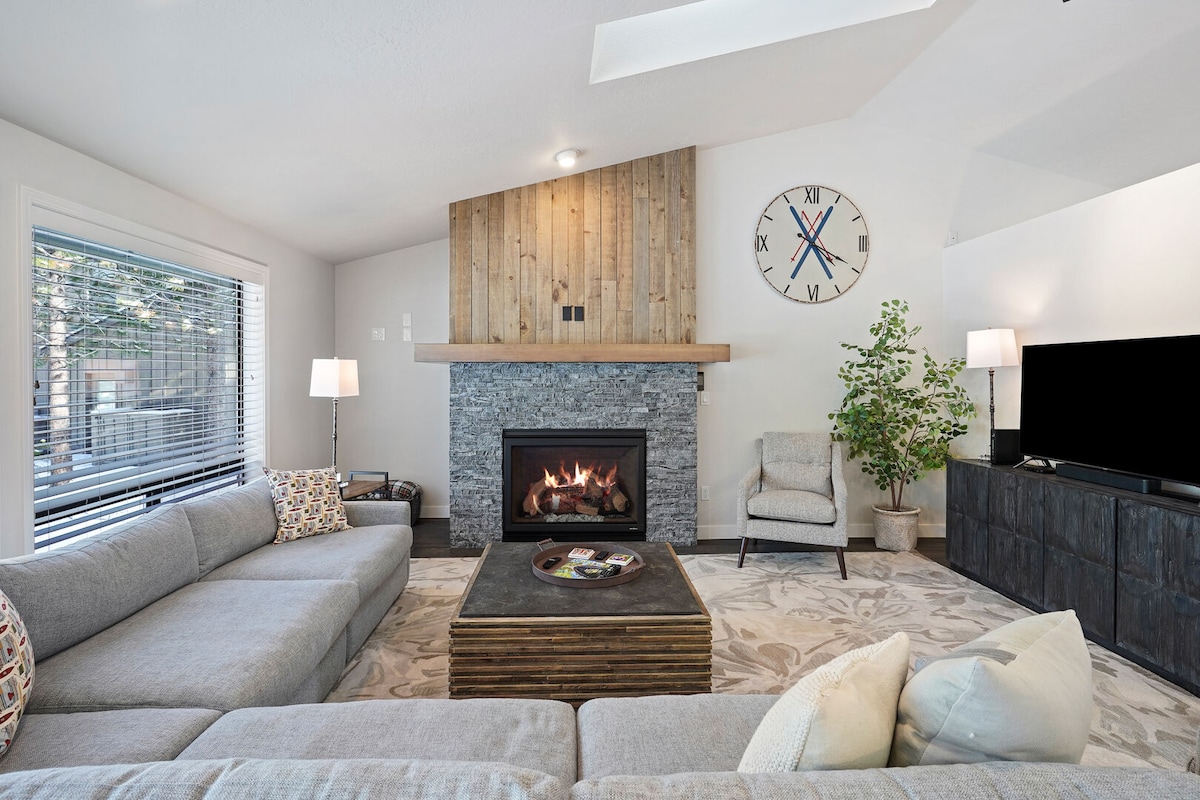 A spacious living room is highlighted by a large sectional sofa and a rustic coffee table. A stone fireplace serves as the focal point, with a wooden mantel above. Natural light fills the space through a window with horizontal blinds, complemented by a green potted plant.