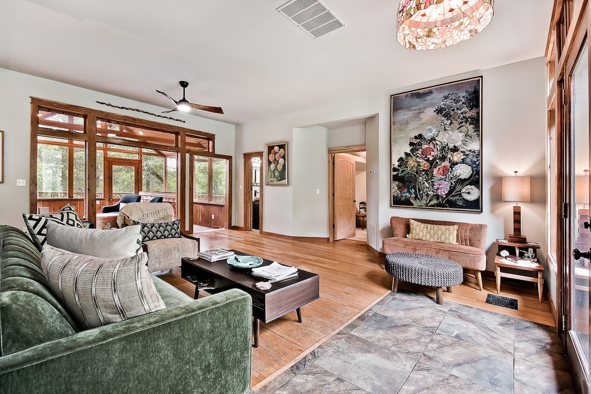 A spacious living area is enhanced by large windows, allowing natural light to fill the room. Comfortable seating includes a green sofa, a cozy armchair, and a stylish ottoman. A colorful floral artwork adorns the wall, complemented by a unique light fixture above.