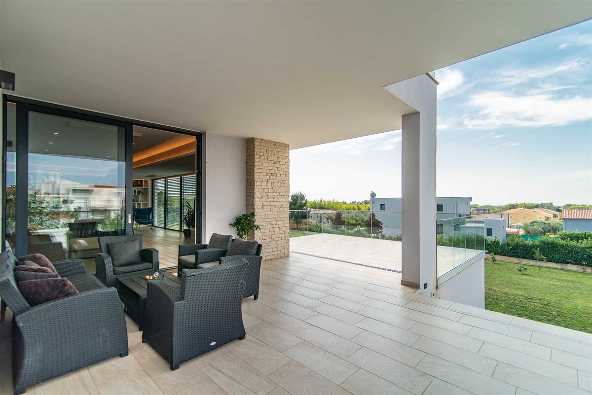 A spacious covered terrace offers comfortable seating arranged around a central table. Large glass doors provide a view into the interior living space, while the terrace extends to an open area with grass and scenic surroundings visible in the distance.